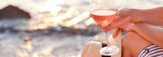 Girl is sitting on sea beach with wineglass of wine at sunset in summer vacation in resort. Tourist woman in striped dress with straw hat is enjoying life, relaxing, drinking, traveling.
