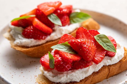 Homemade bruschettas with strawberries, cream cheese and basil in white plate on light background. Summer appetizer. Selective focus.